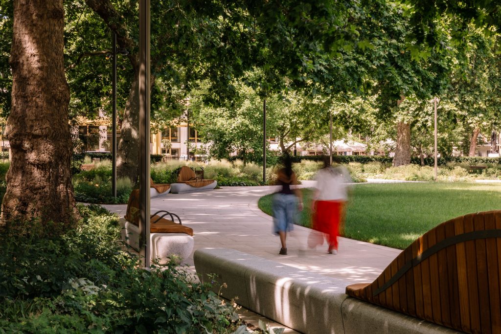 Two people walking through Grosvenor Square. In the foreground is a bench, and you can see the lawn.